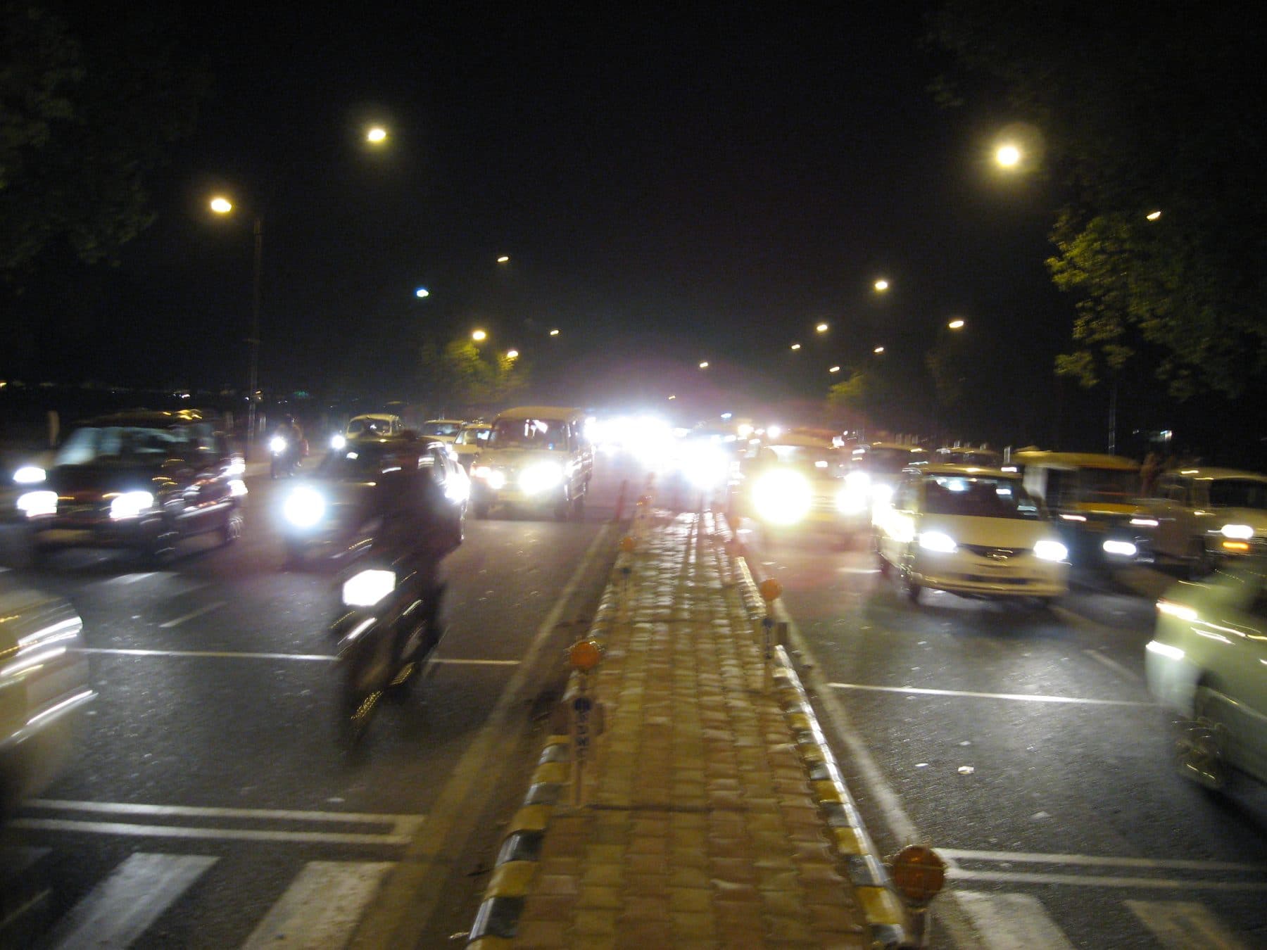 Blurry night-time Delhi street with car headlights, scooters, and streetlamps