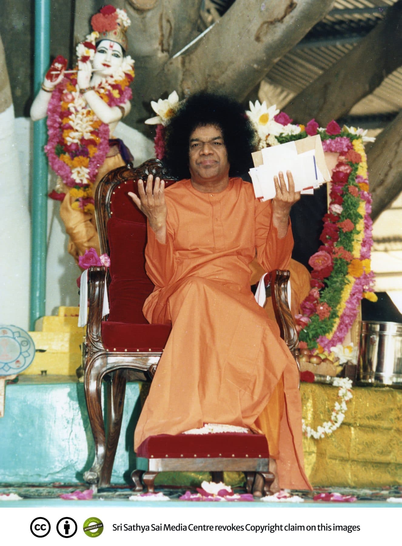 Sathya Sai Baba in orange robe seated on a red chair holding letters at Brindavan ashram, 2010