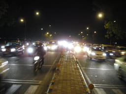 Blurry night-time Delhi street with car headlights, scooters, and streetlamps