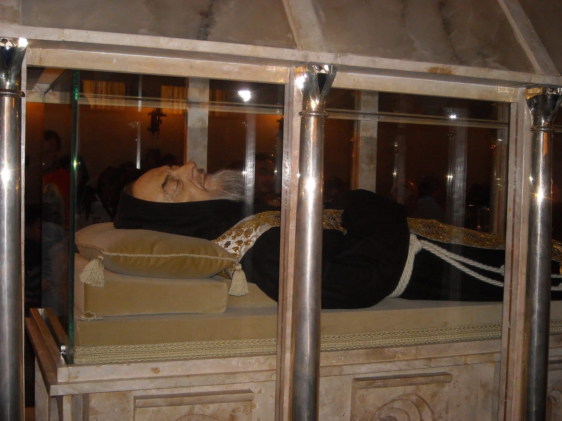 The embalmed body of St Padre Pio in Capuchin habit with clasped hands, lying in a transparent display case at the shrine in San Giovanni Rotondo (the face is a lifelike silicone mask).